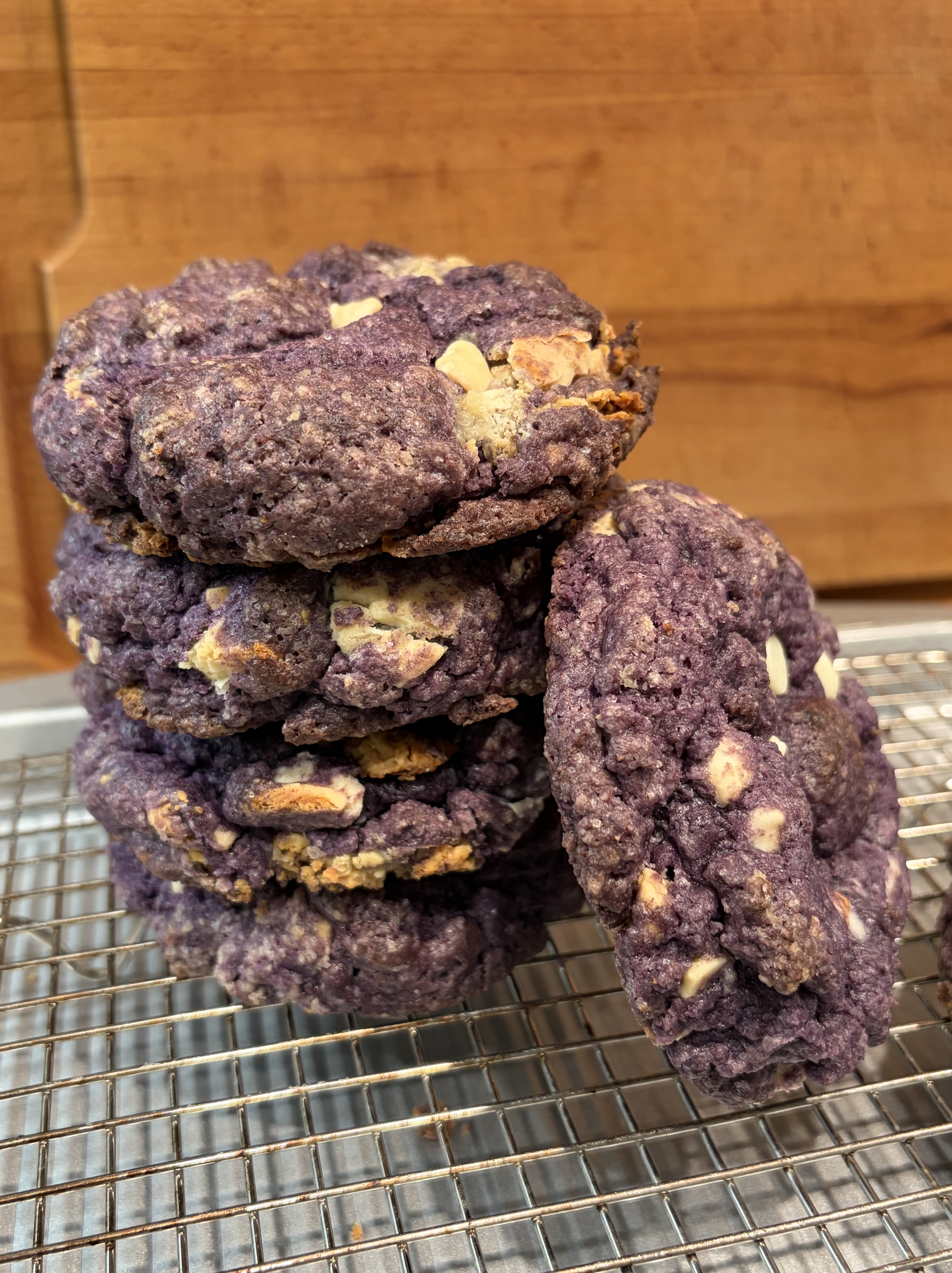 A rustic wooden table with a tray of freshly baked ube sourdough cookies arranged in neat rows. The cookies have crisp golden edges and soft purple centers, with visible cracks that show their chewy texture.